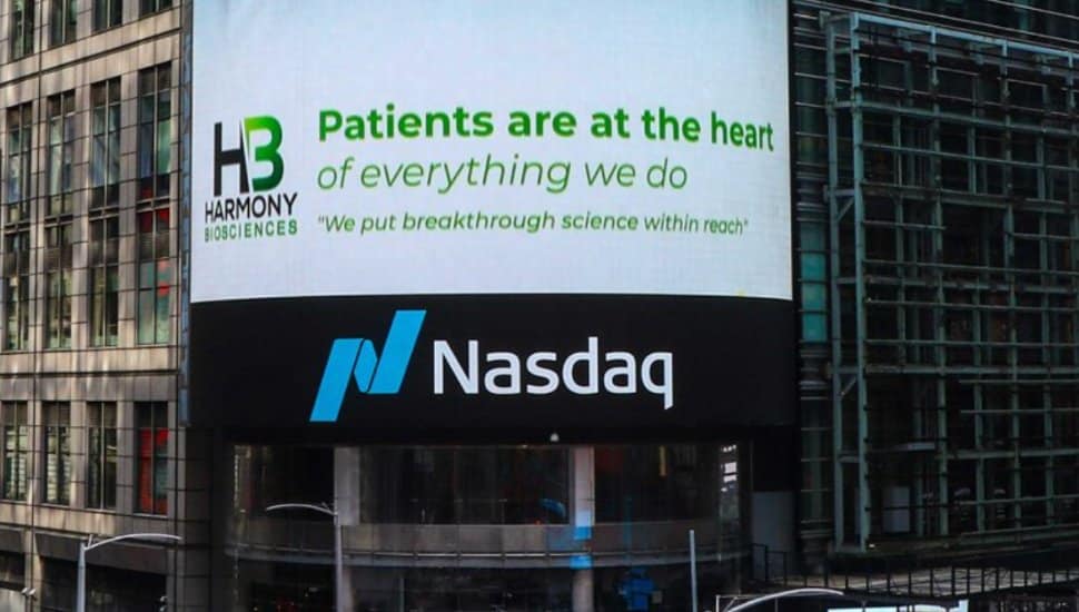 A sign for Harmony Biosciences stands above a sign for NASDAQ on a building in New York.