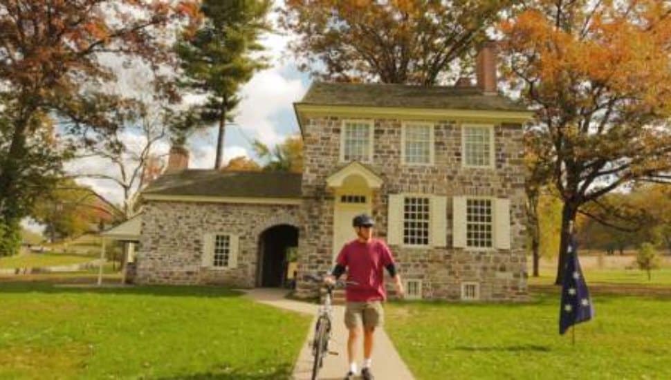 A cyclist walking with his bike in front of a historic stone house at Valley Forge National Historical Park during fall, surrounded by colorful autumn trees.