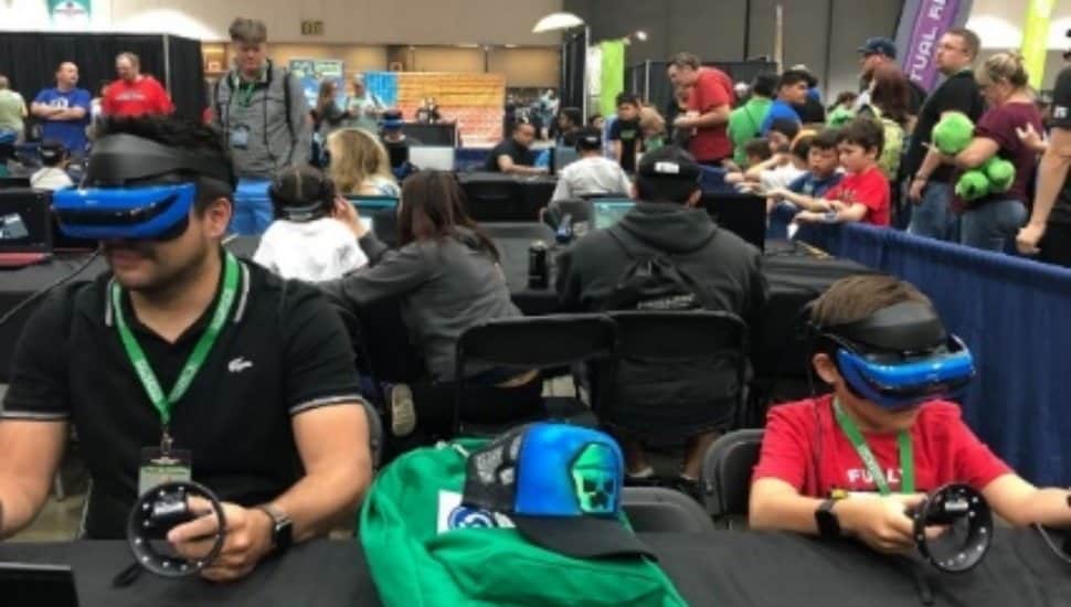 Attendees of all ages wearing VR headsets and using controllers at a gaming station during a family-friendly convention, with kids and parents gathered in the background.