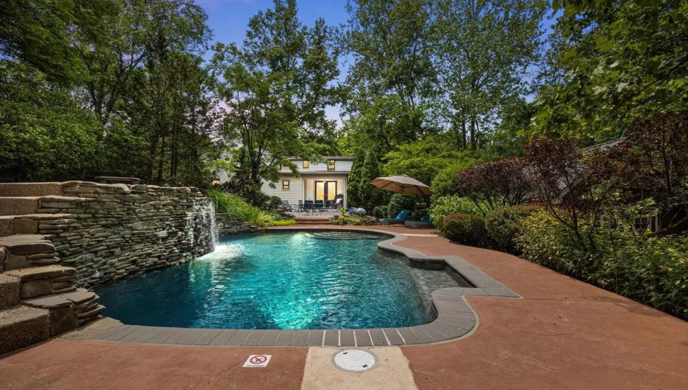 Pool with rock wall surrounded by trees with white stone farmhouse.