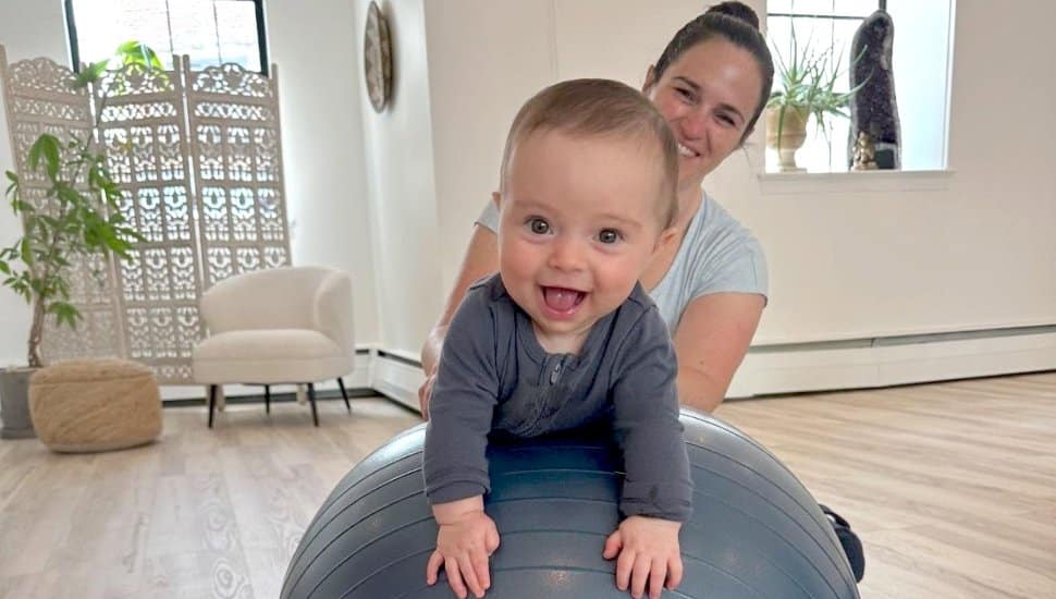 A woman helps her baby on an exercise ball.