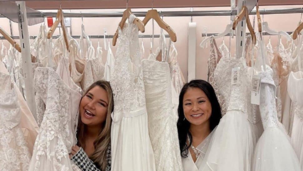 Two women poke their heads out from behind a rack of wedding dresses.