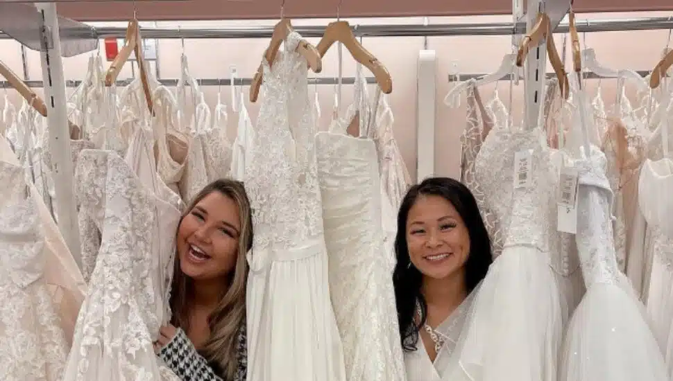 Two women poke their heads out from behind a rack of wedding dresses.