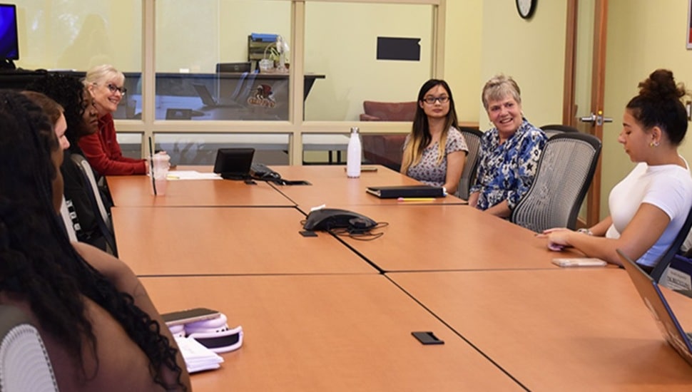 College students and professor have discussion at conference room table.