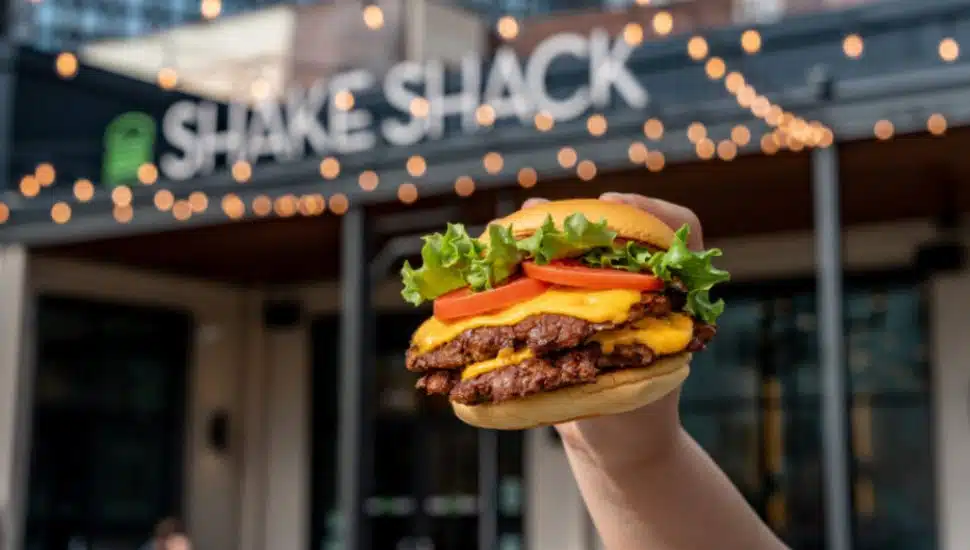 A hand holds a Shack Burger in front of a Shake Shack.