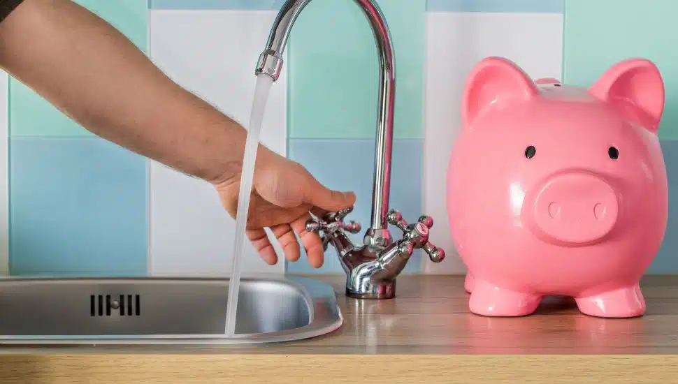 Flowing tap water and a piggy bank standing next to it. Water consumption price concept.