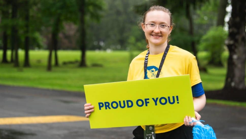 Student wearing a yellow T-shirt smiles while holding a bright green sign that says 'Proud of You!' during an outdoor campus event.