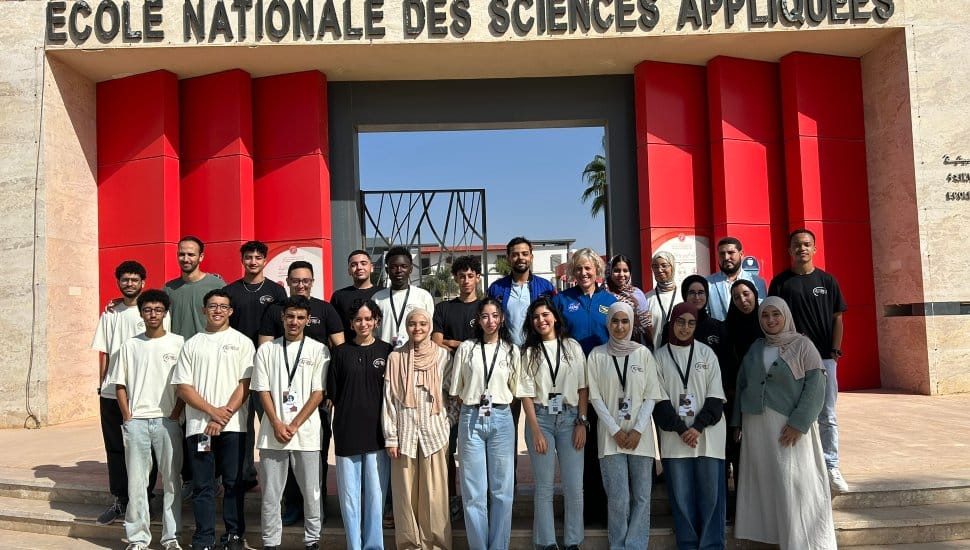 A group of students and faculty stand in front of the entrance to the École Nationale des Sciences Appliquées. The group poses for a photo beneath the school’s sign, with red and beige architectural panels behind them and clear blue sky overhead.