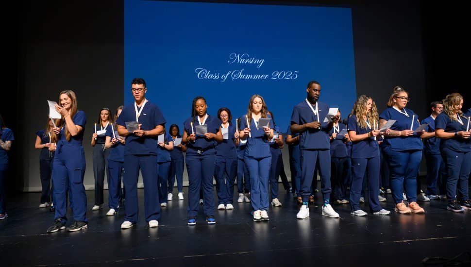 Montgomery County Community College nursing graduates from the Class of Summer 2025 stand on stage in blue scrubs during the pinning ceremony, holding candles and reciting the Nightingale Pledge at the Montco Cultural Center.