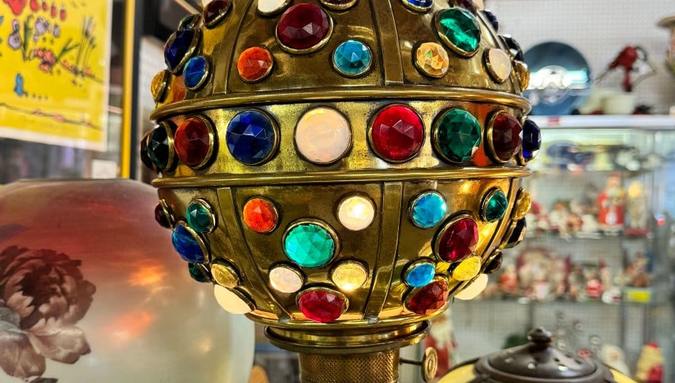 A close-up of a vintage brass lamp adorned with colorful glass gems, displayed among other antique collectibles at a fair.