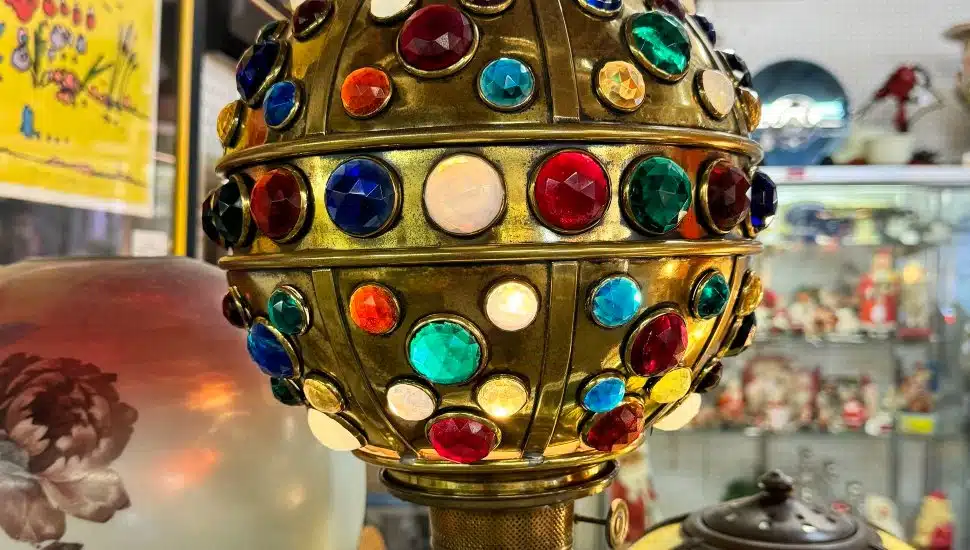 A close-up of a vintage brass lamp adorned with colorful glass gems, displayed among other antique collectibles at a fair.