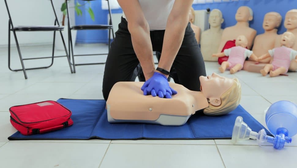 Instructor demonstrating Hands-Only CPR on a training manikin using chest compressions, with AED kit and practice equipment visible during a CPR training session.