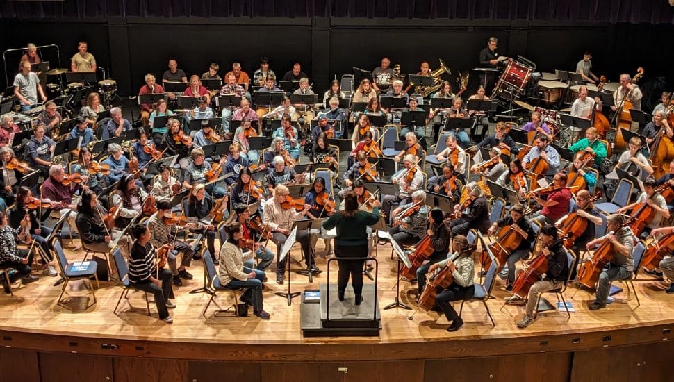 A large symphony orchestra rehearses on stage, with musicians playing strings, brass, woodwinds, and percussion, while a conductor leads from the podium in front.