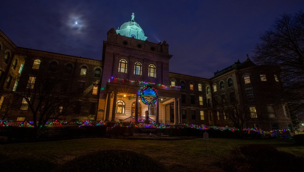 A historic building at Immaculata University is illuminated at night with colorful Christmas lights, featuring a large wreath above the entrance and a glowing dome beneath a moonlit sky.