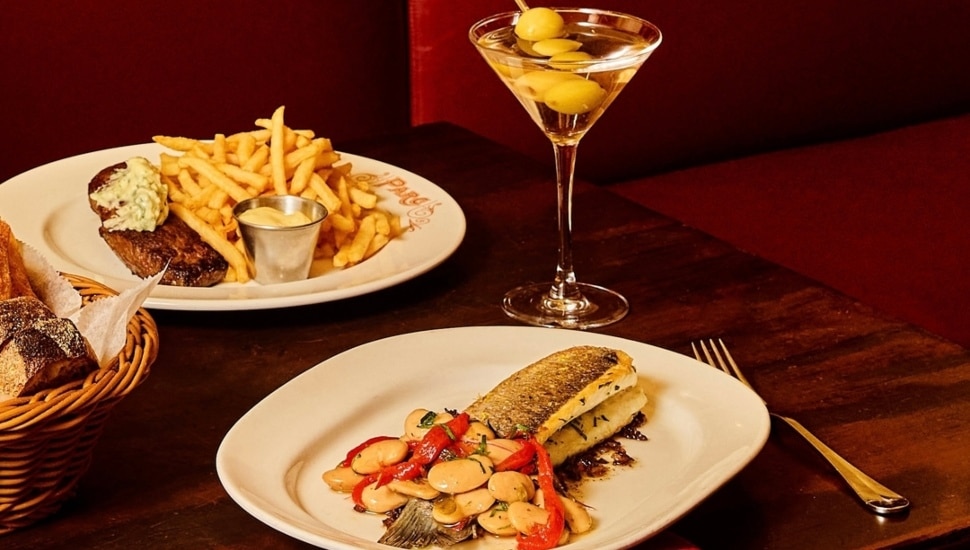 Two plates of food and a martini on a table next to a plate of breadsticks.
