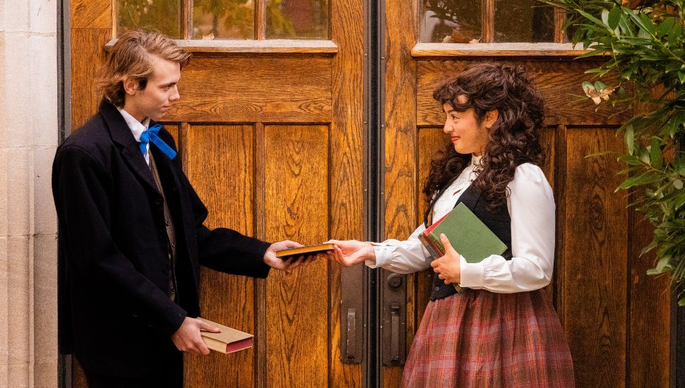 Two actors in period costumes stand in front of large wooden doors, exchanging a small book. The scene evokes a 19th-century setting, reflecting a moment from West Chester University’s production of Little Women, The Broadway Musical.
