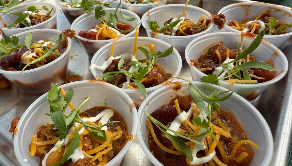 Small plastic cups filled with all-beef chili, topped with shredded cheese, lime crema, and fresh micro cilantro, arranged on a serving tray at a chili cook-off.
