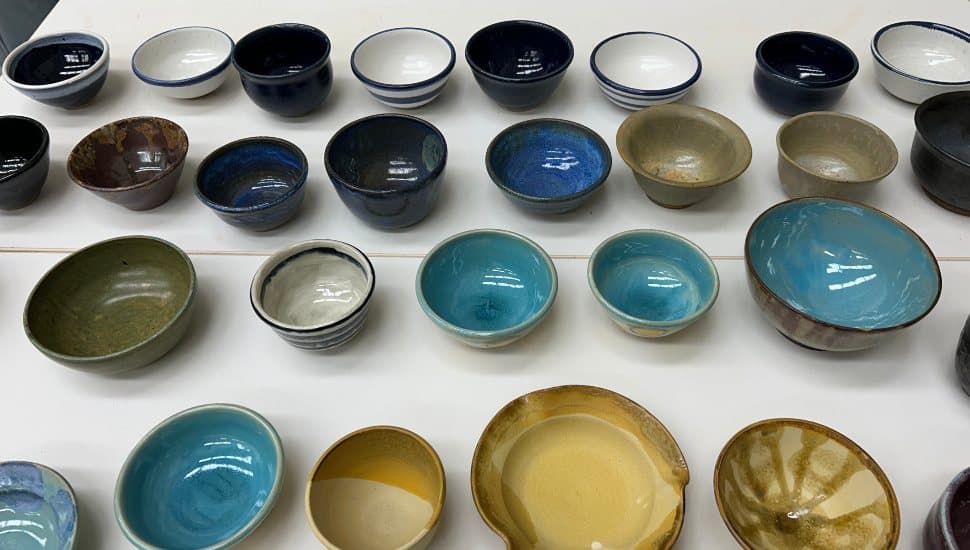 An assortment of handcrafted ceramic bowls in various sizes, shapes, and glazes arranged in rows on a white table. The bowls display a wide range of colors, including blues, greens, browns, and neutrals, showcasing the work of students and faculty for MCCC’s Empty Bowls fundraiser.