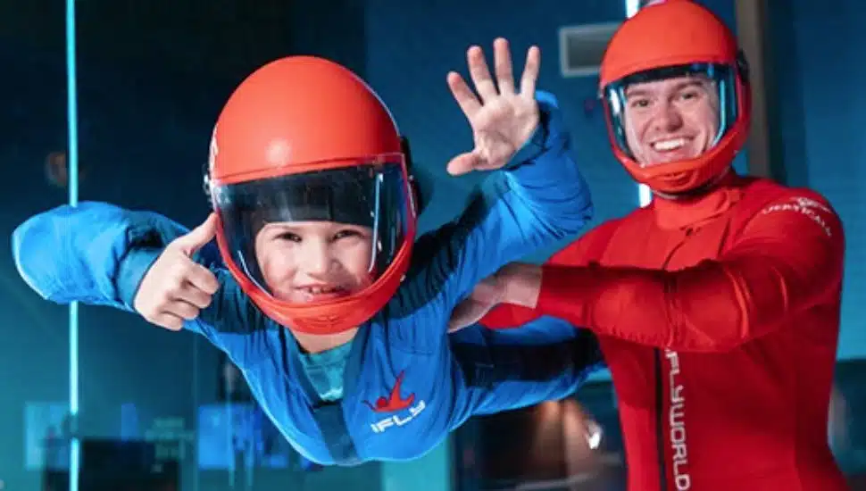 A boy in a flight suit waves as an instructor helps them to balance in the air.