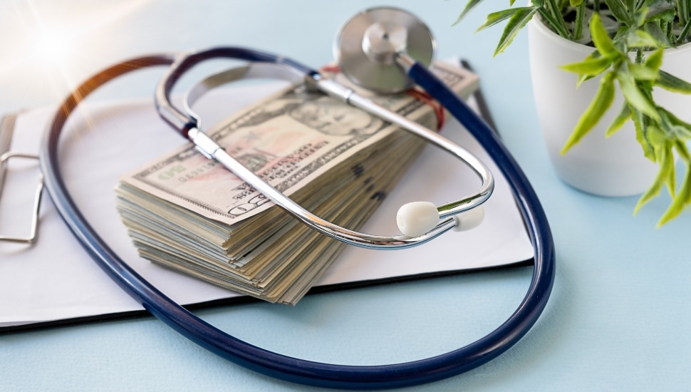 A stethoscope over a stack of money, with a plant in the background at the right corner.