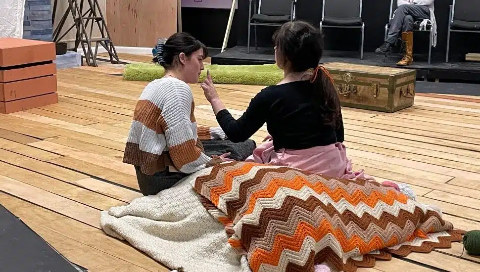 Two actors sit on stage during a theater rehearsal, wrapped in crocheted blankets, while another person observes in the background at Montgomery County Community College.