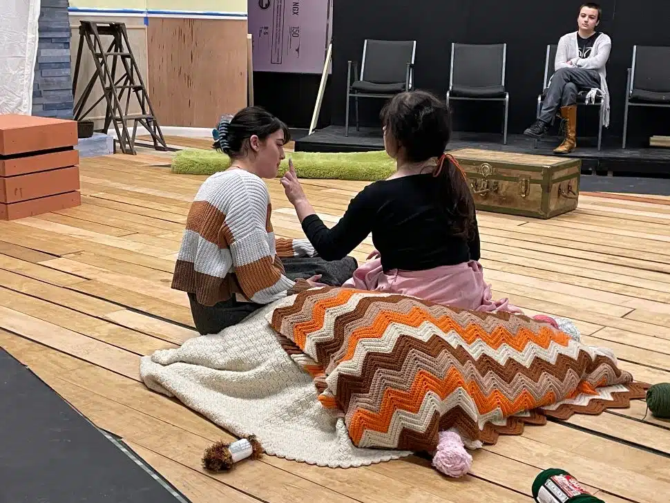 Two actors sit on stage during a theater rehearsal, wrapped in crocheted blankets, while another person observes in the background at Montgomery County Community College.