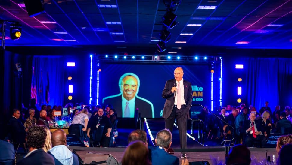 A speaker stands on stage addressing a seated audience at a large event with bright stage lighting and a large projected portrait behind him.