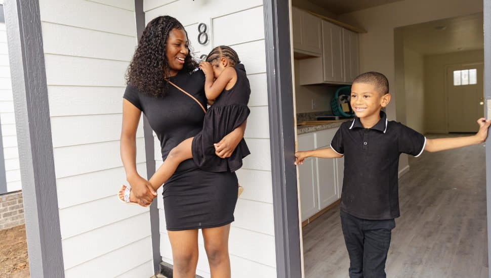 A young mother holds her daughter next to her son outside their new home.