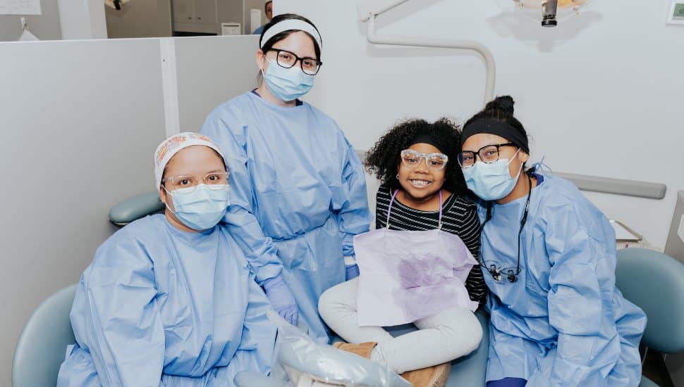 Dental hygiene students and a young patient smiling together at Harcum College’s Sealant Day, wearing protective masks and gowns inside the clinic.