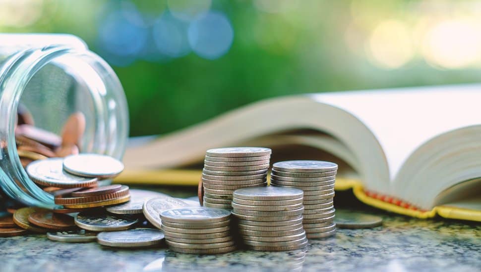 Pile of coins falling out of a jar next to open book for education funding.