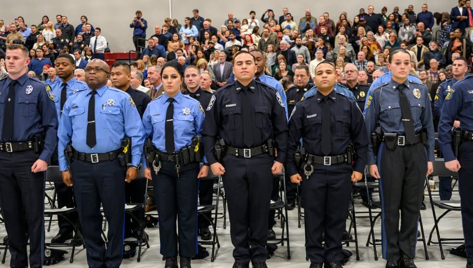 Graduates of the MCCC Municipal Police Academy stand in uniform during a graduation ceremony with an audience behind them.
