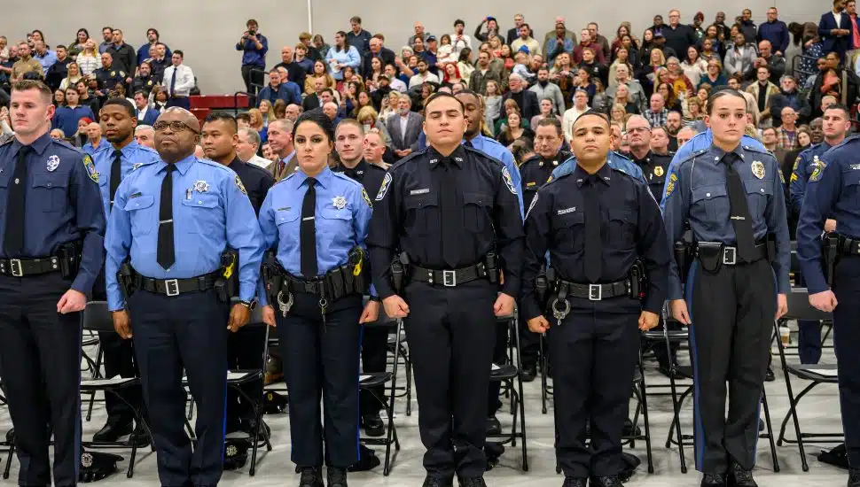 Graduates of the MCCC Municipal Police Academy stand in uniform during a graduation ceremony with an audience behind them.