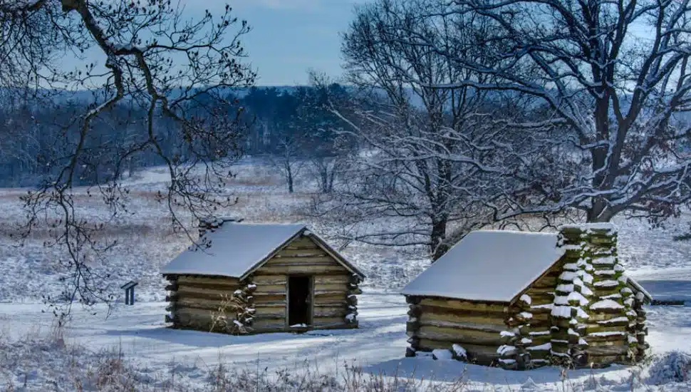 Two historic log cabins in Valley Forge National Historical Park covered in fresh snow, surrounded by bare winter trees and a snowy field.