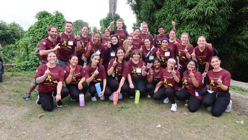 Group of Salus University SOSH students and faculty wearing matching maroon shirts pose together outdoors during a mission trip to Jamaica.