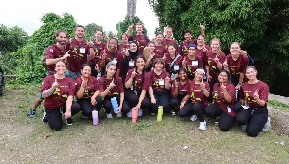 Group of Salus University SOSH students and faculty wearing matching maroon shirts pose together outdoors during a mission trip to Jamaica.