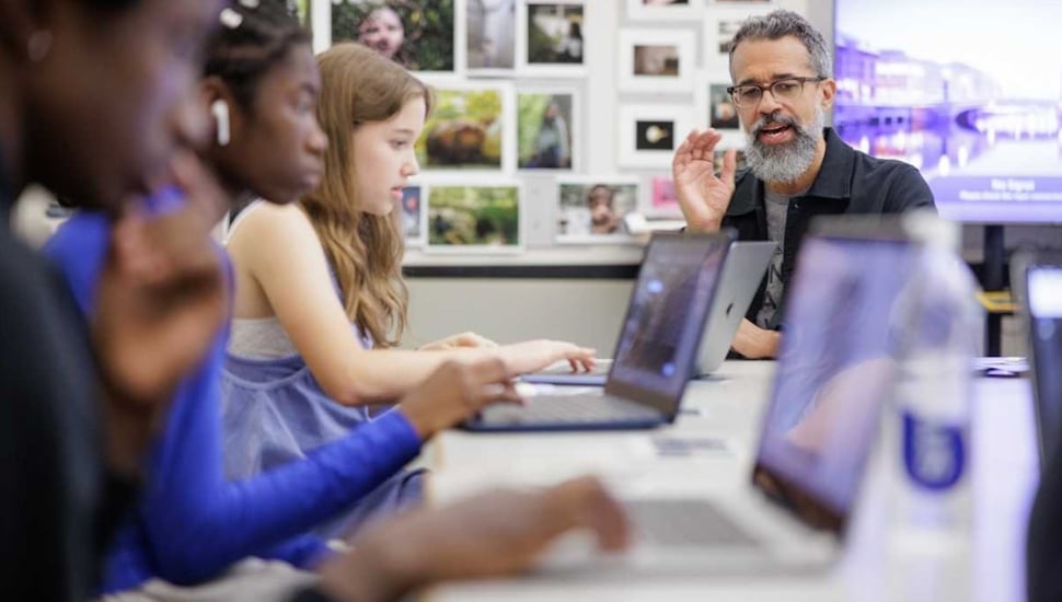 Students learn on laptops with teacher in background.