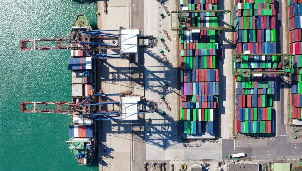 An aerial view depicts a harbor dock with ship freight stacked beside each other.