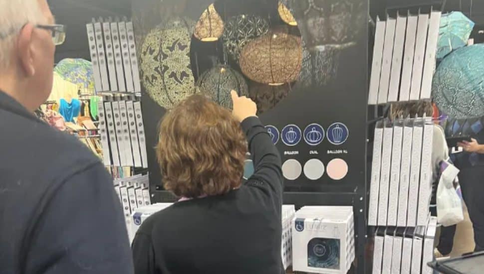 Two attendees examine decorative hanging lanterns at a gift show booth, with one person pointing toward a display of patterned globe lights.