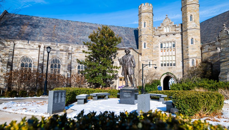 Snow-covered West Chester University campus featuring the Frederick Douglass statue in the foreground, with historic stone academic buildings and towers under a bright blue winter sky.