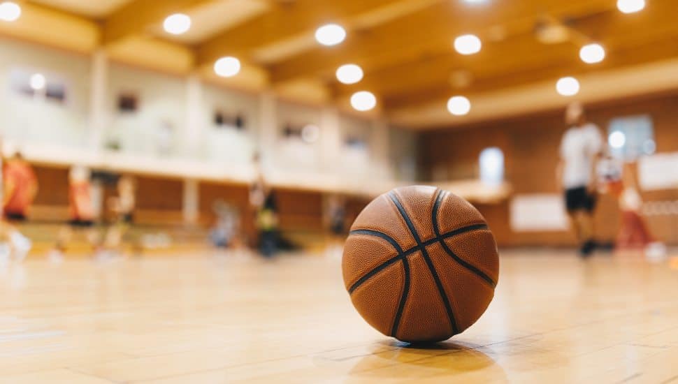 Basketball on Wooden Court Floor Close Up with Blurred Players Playing Basketball Game in the Background.