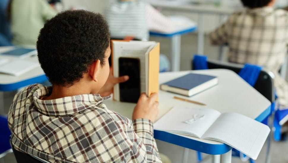 Back view at young African American kid hiding smartphone in book and cheating in class.