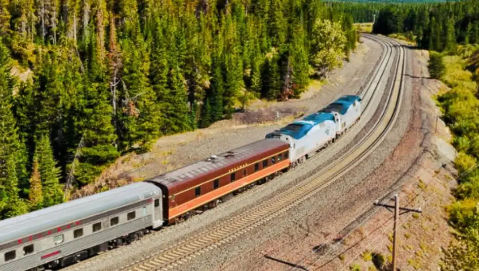 An AMTRAK train with a Lunatrain cart rides through the countryside.