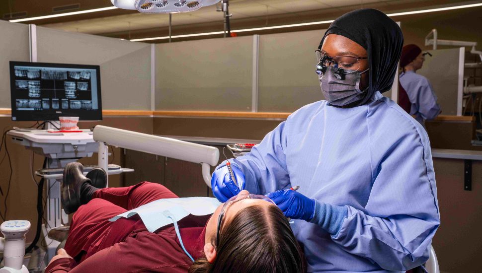 Dental hygiene student wearing protective gear and magnifying loupes provides oral care to a patient in a clinical setting under supervision.