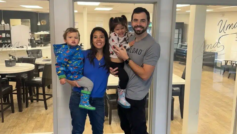 A smiling family of four stands in front of a café entrance with glass doors. A woman in a blue top holds a young boy wearing a colorful jacket and rain boots, while a man in a gray T-shirt holds a little girl with pigtails and rain boots. The café interior with tables and chairs is visible behind them.