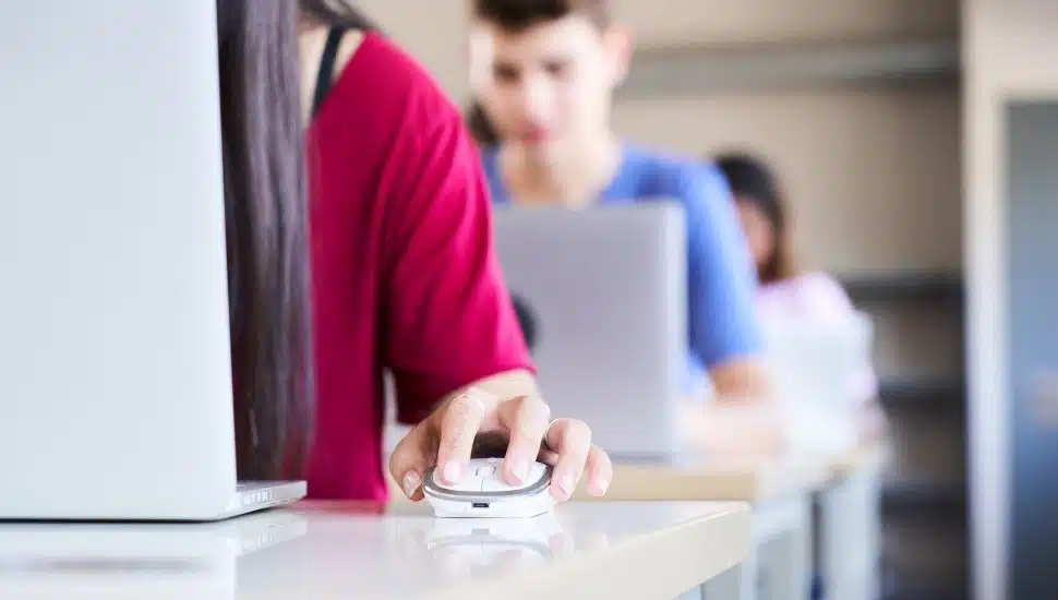 Close-up of a hand clutching a mouse in a high school classroom, a group of cheerful students study with a laptop computer.