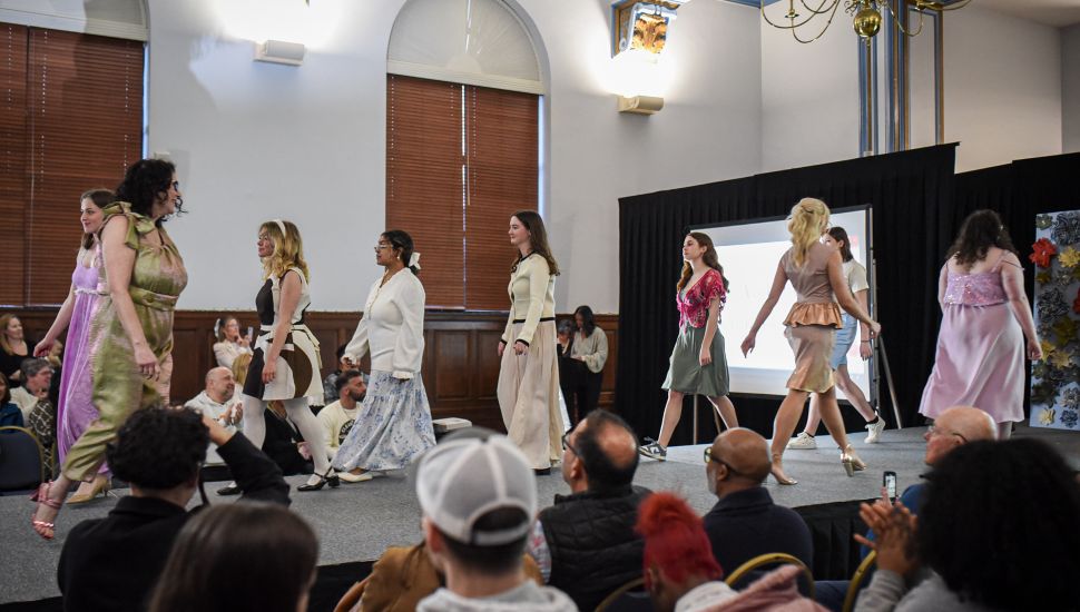 Fashion merchandising students and models walk the runway during the Immaculata University IU Fashion Show in the Great Hall as audience members watch and applaud.