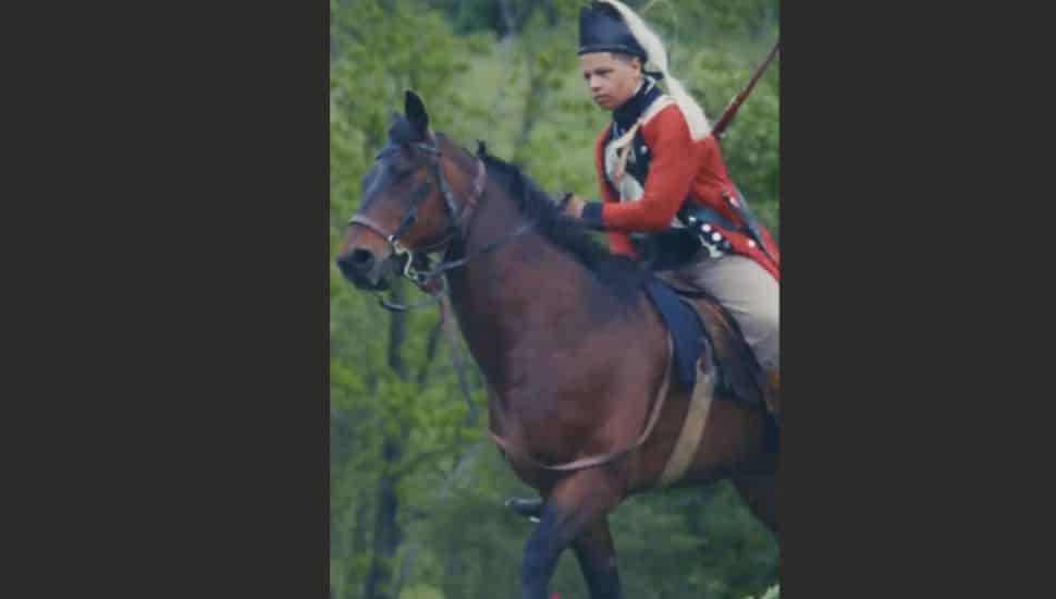 Historical reenactor dressed in a Revolutionary War uniform riding a brown horse outdoors.