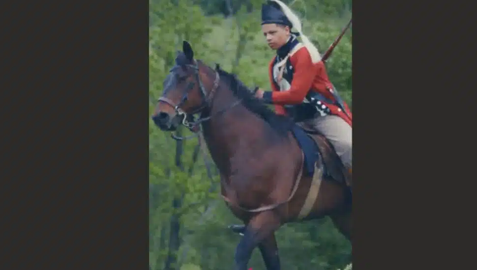 Historical reenactor dressed in a Revolutionary War uniform riding a brown horse outdoors.