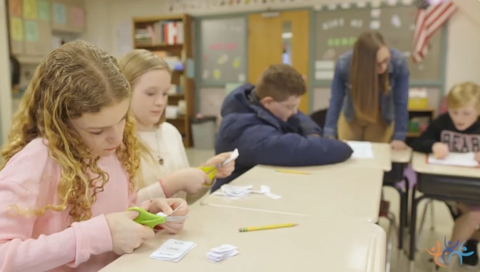 Boys and girls sit at a table working on a project with a teacher in the background.