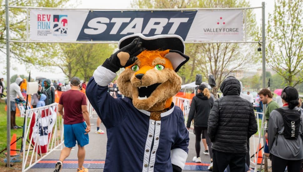 A costumed mascot waves at the start line of the Valley Forge Revolutionary Run as participants gather behind at a spring race event.